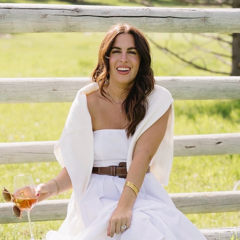 A woman in a white, strapless dress sits outdoors on grass, smiling and holding a glass of rosé wine. She has long brown hair and wears a white sweater draped over her shoulders and gold bracelets on her wrist.