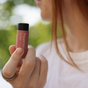 A person with long hair holds a tube of Hearth and Homestead Tallow Lip Balm close to their face, with blurred green foliage in the background.