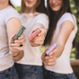 Three women in white shirts hold out colorful tubes of Hearth and Homestead Tallow Lip Balm toward the camera, their faces slightly out of focus with greenery in the background.