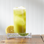 A tall glass of iced green tea sits on a wooden surface with a lemon slice beside it. In front is a packet labeled The Republic of Tea Organic Matcha Single Sips Tea.