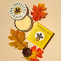 A NOPALERA Moisturizing Botanical Bar in a round tin and its yellow box rest on a beige background with autumn leaves and a pine cone, showcasing this sustainable, botanical moisturizer.