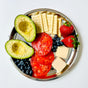 The Ahimsa Purposeful Plate, a round stainless steel dinner plate, is shown on a white background holding avocado slices, tomato slices, rectangular crackers, strawberries, blueberries, and two cheese cubes.