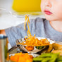 A child in a gray shirt eats spaghetti from the Ahimsa Smart Snacking Bowl—a stainless steel, plastic-free option that keeps meals healthy with noodles, green beans, and orange vegetables.