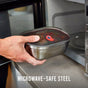 A hand places a Black + Blum Multi-Function Food Box Mixed Set (1.2L, stainless steel with plastic lid) into a microwave. Text on image: MICROWAVE-SAFE STEEL 1.2L BOXES.