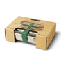 The Black + Blum Stainless Steel Lunch Box Large is leak-proof, secured with a green silicone strap, and comes partially enclosed in a brown cardboard box featuring product information and illustrations.