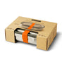 The Black + Blum Stainless Steel Lunch Box Large is leak-proof, secured with an orange elastic band, and comes partially enclosed in a brown cardboard sleeve displaying product details and the brand name.