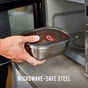 A hand puts the Black + Blum Steel Food Box with a plastic lid into a microwave. “MICROWAVE-SAFE STEEL” appears below, emphasizing this leakproof lunch box as a smart food storage choice.