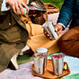 Two people enjoy a picnic as one pours a drink from a bottle into a Black + Blum Steel Cup Set of 4; three more cups, two with ice and lime, rest on a bamboo chopping board nearby.