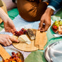 A close-up of hands reaching for cheese, grapes, crackers, and a goat cheese log on a bamboo board during a picnic. Black + Blum's Steel Plate Set of 4 and more food rest on a green-and-white blanket in the background.