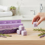 A hand picks up a Soapymania Lavender Tea Sugar Scrub Cube from a stack on wood, with the scrub box, rosemary sprigs, and lavender flowers beside it—ideal for exfoliation and skincare rituals in your bathroom.
