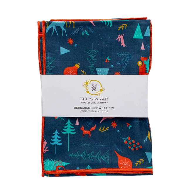 A folded Bees Wrap Reusable Gift Wrap made from organic cotton, features dark blue fabric with colorful woodland animals and trees, wrapped in a white paper band with the Bees Wrap logo and product details.
