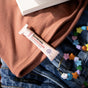 A tube of Sunniemade Nourishing Lip Balm - Blossom Butter rests on a folded brown shirt, next to blue jeans and a colorful beaded necklace with star-shaped beads.