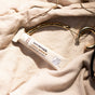A tube of Sunniemade Hydrating Lip Balm - Coco Quench sits atop a soft beige fabric beside a set of gold hoop earrings. Labeled Coconut & Vanilla and made with nourishing cocoa butter, this lip balm features sleek, recyclable packaging.
