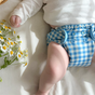 A baby in a white long-sleeve top and an Esembly Baby Outer Diaper Cover + Swim Diaper, Size 1 (100% OEKO-TEX Certified Recycled Fabric), lies on a light surface beside fresh chamomile flowers.