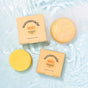 Two Sunniemade Moisturizing Conditioner Bars - Mighty Mango and two matching boxes rest on a light blue, rippling water surface. The round, yellow-orange bars are enriched with mango butter for gentle conditioning, while the peach-colored boxes—printed with product details—highlight their plastic free packaging.
