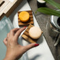 A hand with red nail polish reaches for a round, beige soap bar—infused with hydrating mango butter—on a wooden soap dish next to a Sunniemade Moisturizing Shampoo Bar - Mighty Mango in yellow, with a plant and metal kettle nearby by the bathtub.