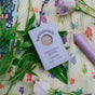 A box of Sunniemade Exfoliating Body Bar Soap - Purple Rain, enriched with Brazilian purple clay, rests on a floral cloth alongside green leaves, a purple candle, a light purple flower, and a floral notebook.