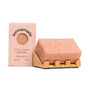 A pink Exfoliating Body Bar Soap – Rise & Grind by Sunniemade, speckled with black coffee grounds for gentle exfoliation, sits on a wooden soap dish. Beside it is the matching Sunniemade box labeled “Rise & Grind,” complete with a round window displaying the soap inside—a perfect addition to your natural body care routine.