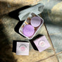 Two round purple Sunniemade Strengthening Shampoo Bars—Super Bloom—rest in a white container, bathed in sunlight that casts gentle shadows. Beside the container on a stone surface are two light purple Sunniemade boxes labeled Super Bloom and High Noon, each featuring plastic-free shampoo bars formulated with nourishing rosehip oil.