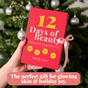 A hand holds a red FaceTory 12 Days of Beauty Sheet Mask Set gift box with a pink ribbon in front of a decorated Christmas tree. Text below reads: “The perfect gift for all skin types & holiday joy.”.