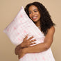 A smiling woman hugs the Kitsch Standard Satin Pillowcase with a light pink cover featuring thin red lines and bows, standing before a beige background—ideal for reducing hair breakage and controlling frizz.