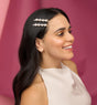 A woman with long dark hair smiles, styling her hair with Kitsch Metal Gemstone Bobby Pins and pearl clips. She wears gold eyeshadow, a sleeveless beige top, and hoop earrings against a pink background.
