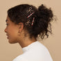 A woman with curly dark hair in a loose bun, wearing gold hoop earrings and a white ribbed top, accents her look with Kitsch Metal Gemstone and Bow Bobby Pins—gold bows and red-and-white gemstones styled on the side of her hair.