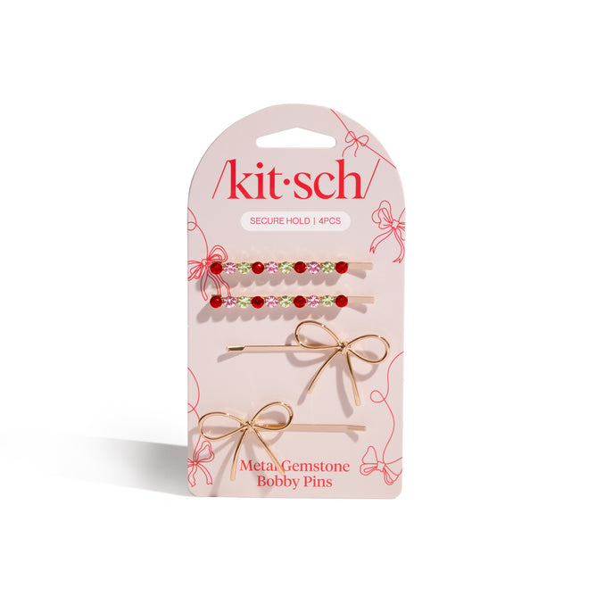 The Kitsch Metal Gemstone and Bow Bobby Pins set includes four playful bobby pins—two with colorful gemstones and two gold bows—presented on a pink card with red bow outlines.