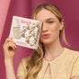 A woman with long blonde hair holds a Kitsch Happy Holiday Hair Set box by her face, wearing a light gold top against a pink background to showcase the festive satin scrunches inside.