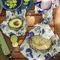 A wooden table displays a halved avocado, sliced limes, tortillas, red onion, and chopped avocado, partly wrapped in Bees Wrap Lunch Pack by Bees Wrap—an eco-friendly alternative with blue animal patterns. A knife and cutting board are also visible.