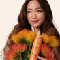 A woman with long, wavy brown hair holds a bouquet of orange-yellow flowers and an orange bottle of The Powder Shampoo Strengthening & Soothing Powder Shampoo, smiling gently as she looks to the side against a neutral background.