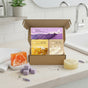 A Soapymania Body Care Set with exfoliating bar soap, scrub, and loofah sits open on the counter. Next to it are an orange bar soap, purple sugar scrub cubes, a round yellow soap, a white towel, and a sink with tiled backsplash.