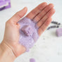 A hand holds a purple Soapymania Lavender Tea Sugar Scrub Cube with bubbles in the palm. In the background, more scrub cubes and lavender sprigs sit on a white surface.