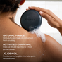 A person uses the Ember Wellness Charcoal Exfoliating Cleanser Bar, a black bar, on their shoulder. Text highlights natural pumice for gentle exfoliation, activated charcoal for detoxifying the skin, and jojoba oil for nourishing hydration.