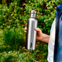 A person holds a Black + Blum Insulated Water Bottle with a brown strap outdoors, surrounded by green foliage. Only their hand and blue jacket are visible, showcasing how this bottle keeps drinks cold.
