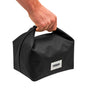 A hand holds the handle of a Black + Blum Lunch Bag, a black rectangular bag with a small white label on the front, perfect for carrying lunch or snacks, shown against a white background.