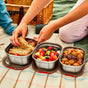 A person arranges sandwiches, fruit, and snacks in a Black + Blum Multi-Function Food Box Mixed Set—some 1.2L—on a picnic blanket, with a wicker basket and drinks nearby.