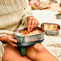 A person in a cream knit sweater sits outdoors on a picnic blanket holding the Black + Blum Stainless Steel Lunch Box Large, filled with pretzels, sandwiches, and snacks. Another container with a sandwich appears in the background.