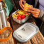 Sitting outdoors on a wooden bench, a person holds a sandwich over the Black + Blum Stainless Steel Lunch Box Large, filled with cherry tomatoes, grapes, and lettuce. The lunchbox lid and a stainless steel water bottle are nearby.