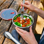 Someone enjoys a salad of leafy greens, roasted veggies, and feta in the Black + Blum Steel Food Bowl at a rustic wooden picnic table, using a fork with leakproof food storage utensils nearby.