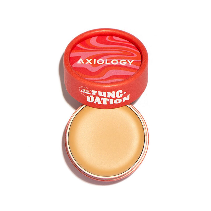 A red circular container of Axiology Fungdation - Mushroom Foundation with a partially open lid showcases the solid beige balm inside. The lid displays bold, wavy patterns and white text.