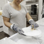 Wearing Full Circle's High & Dry Cleaning Gloves, a person smiles while washing a white plate with a brush under running water in a modern kitchen sink.