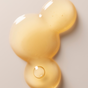 A golden, translucent blob with three connected circles and a small bubble inside, resembling Three Ships Jelly Drops Kakadu Plum + Mushroom Hyaluronic Acid Serum, rests on a smooth, light surface.