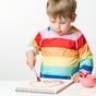 A young child in a colorful striped hoodie uses the Elseware Unplug Paintbrush Set with water-based paints to create a red face in a sketchbook, focusing intently beside a pink bowl of paint on the white table.