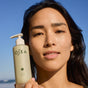 A woman with long dark hair holds up a bottle of OSEA Ocean Cleanser in front of her face, standing outdoors against a clear blue sky.
