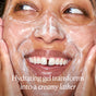 A close-up of a smiling person washing their face with OSEA Ocean Cleanser, covered in a creamy lather. Text reads: This pH-balanced gel face wash transforms into a rich, hydrating foam.