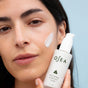 A person with dark hair applies a white cream to their cheek while holding a bottle of OSEA Atmosphere Protection® Cream, a seaweed-based moisturizer, against a light blue background.