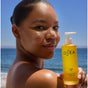 A person smiles at the camera, holding a bottle of OSEA Undaria Algae® Body Oil by the ocean, with sunlight illuminating their glowing, visibly elastic skin.