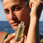 Standing outdoors by the ocean in natural light, a person uses a dropper to apply OSEA Hyaluronic Acid Sea Serum to their face, helping improve skin firmness.
