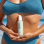A person in a blue textured bikini stands on the beach holding OSEA Hyaluronic Body Serum—an ultra-hydrating treatment ideal for sensitive skin. The ocean and sky are softly blurred in the background.